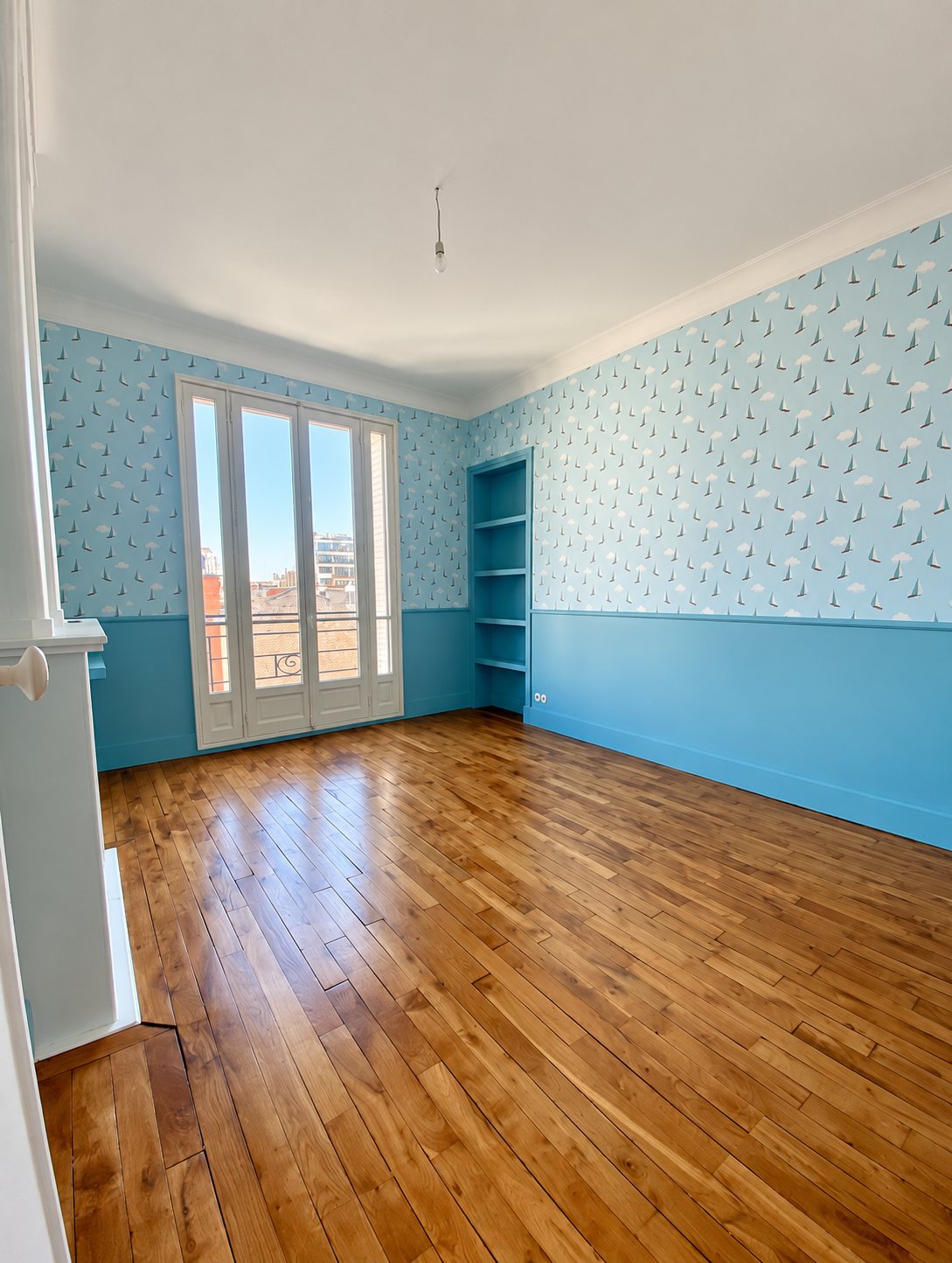 Pièce vide fraîchement rénovée avec papier peint bleu à motifs de voiliers, soubassement bleu turquoise, étagères intégrées, porte-fenêtre et parquet en bois verni.