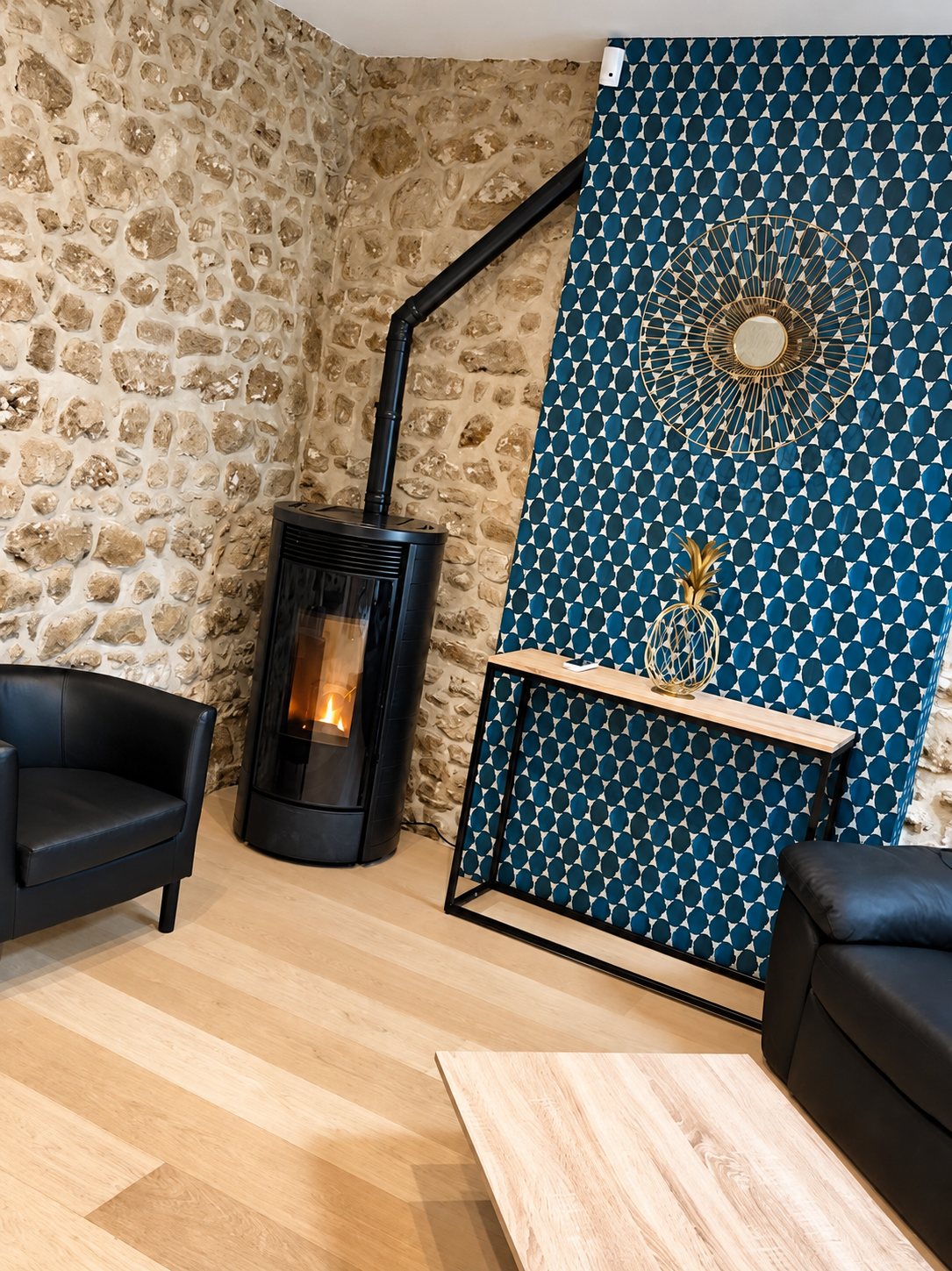 Salon rénové avec un poêle noir dans un coin de mur en pierres apparentes, un mur à motifs bleus orné d’un miroir doré, une console et des fauteuils en cuir noir.
